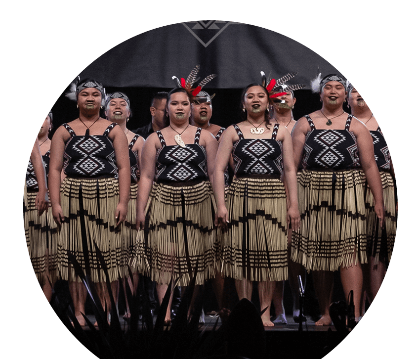 A kapa haka rōpū performs on stage, proudly adorned in black, white, and red kākahu - including traditional piu piu and a feathered Tīpare.
