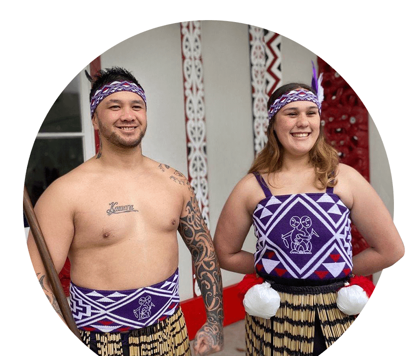 A proud Tāne and Wahine standing in front of a marae, dressed in custom-knitted purple, white and red Kākahu -made by Kapa Haka Makers.