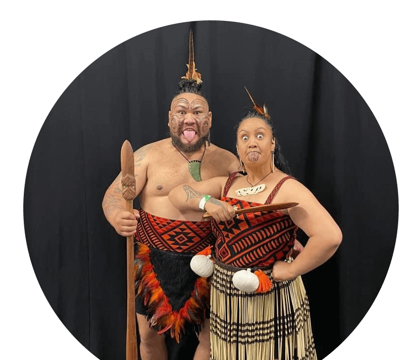 A Tāne and Wahine, dressed in traditional Māori kākahu in bold red and black, channel strength and intensity through a powerful pūkana.
